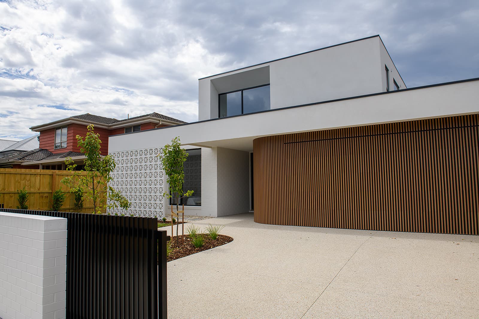 Street facade of Patricia in Keilor East: a white rendered upper form floating over a curved timber batten garage and breeze block screen